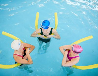 Above view at group of active senior women working out in swimming pool holding pool noodles, copy space